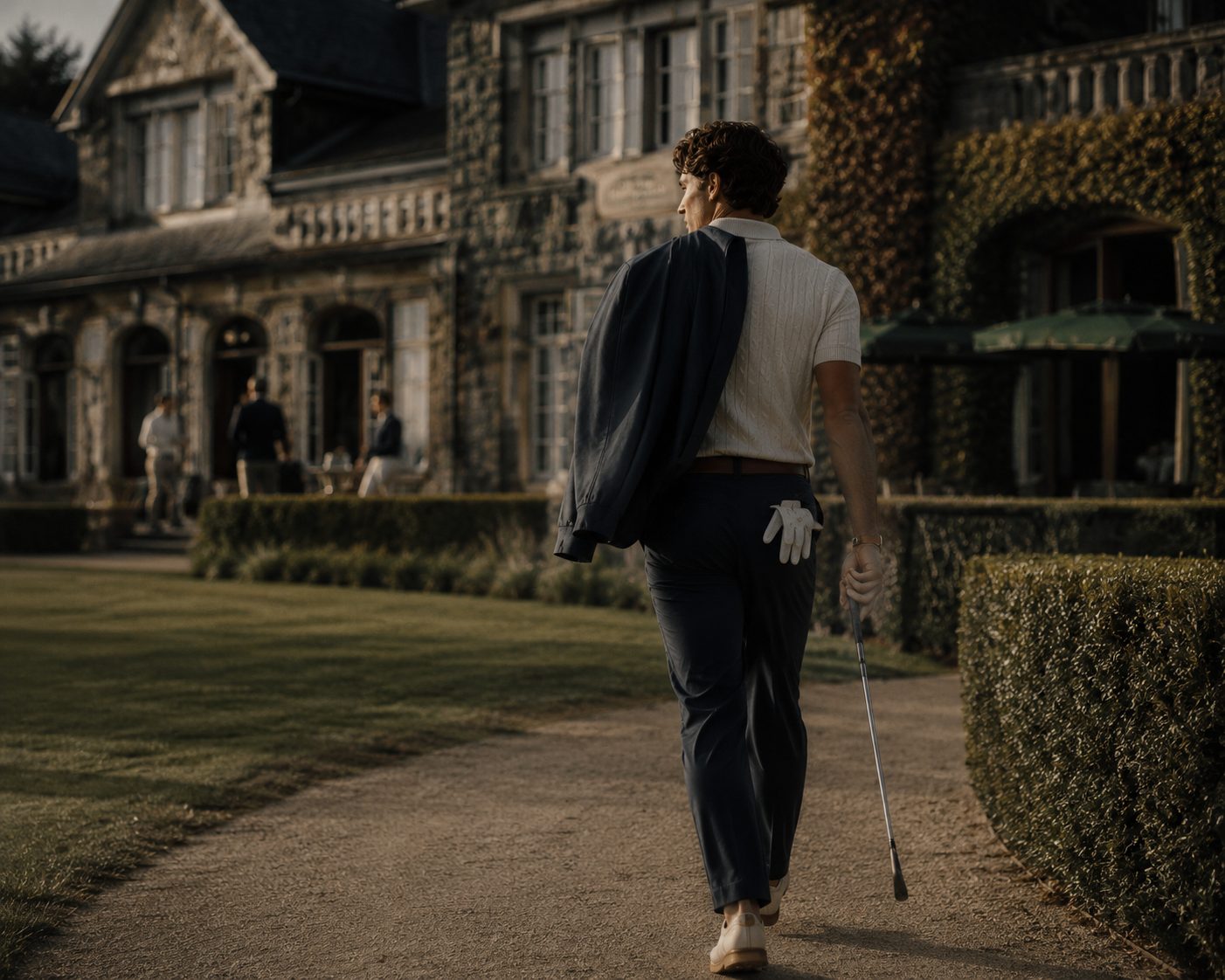 RLX golfer walking toward a stone heritage clubhouse in soft morning light, jacket over the shoulder and club in hand.